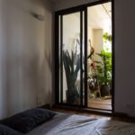 Spálňa s drevenou podlahou, sivou posteľnou bielizňou a čiernymi posuvnými dverami na balkón so zeleňou.