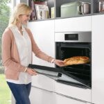 Biela moderná kuchyňa so zabudovanou nerezovou rúrou, z ktorej žena vyberá upečený chlieb.