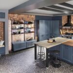 Moderná tmavomodrá kuchyňa v industriálnom štýle s tehlovou stenou, otvorenými regálmi a tmavou mozaikovou dlažbou.