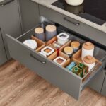 Sivá kuchynská skrinka s otvorenou zásuvkou, kde sú uložené nádoby s potravinami v drevených a bielych organizéroch.