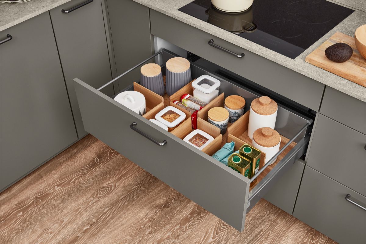 Sivá kuchynská skrinka s otvorenou zásuvkou, kde sú uložené nádoby s potravinami v drevených a bielych organizéroch.