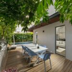 Vonkajšia drevená terasa s bielym jedálenským stolom a modrými stoličkami, tienená stromami pri bazéne.