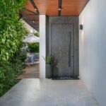 Vchod do domu s betónovými schodmi, tmavými dverami a dreveným podhľadom,.