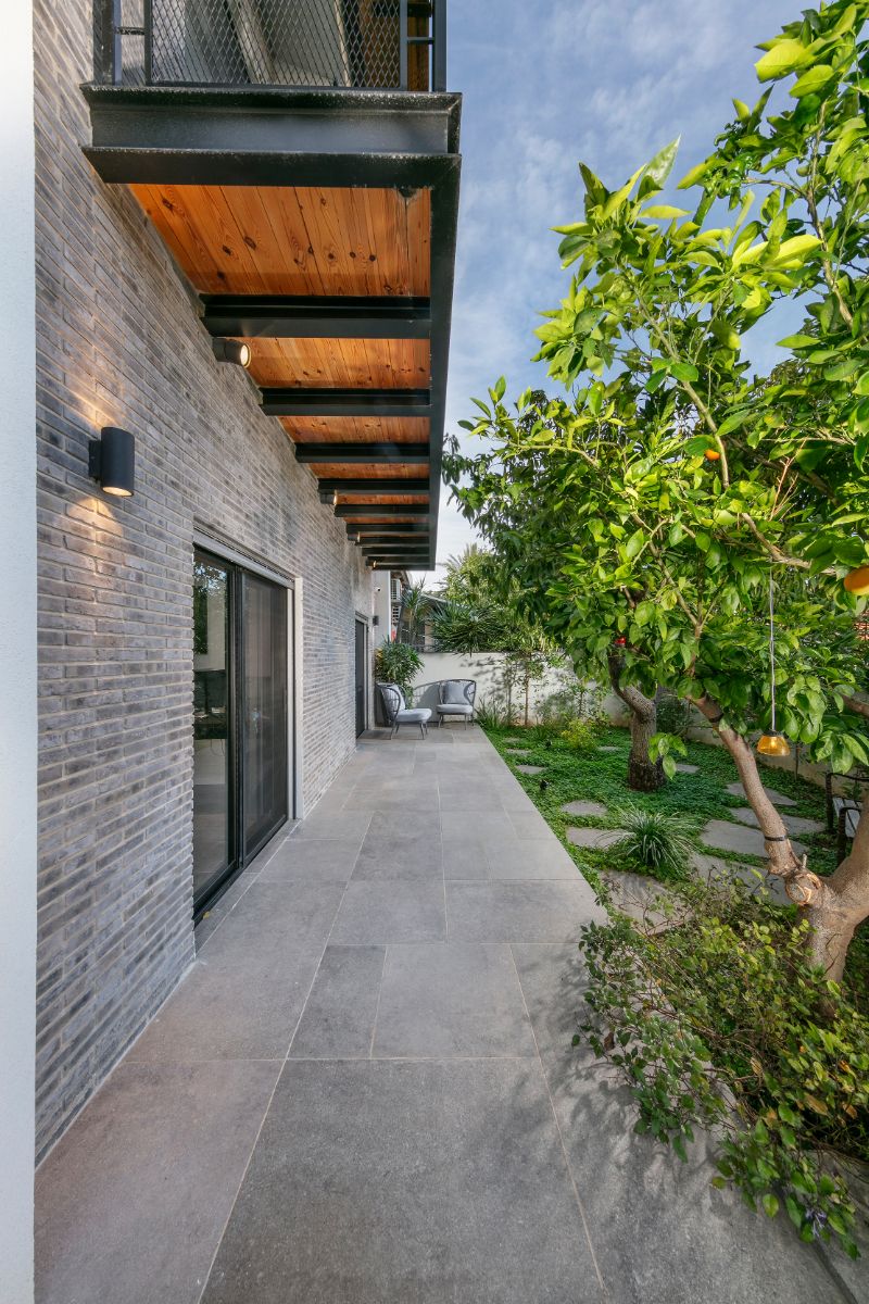 Fasáda domu z tmavých tehál so šedou kamennou dlažbou, tienená dreveným presahom a ovocnými stromami.