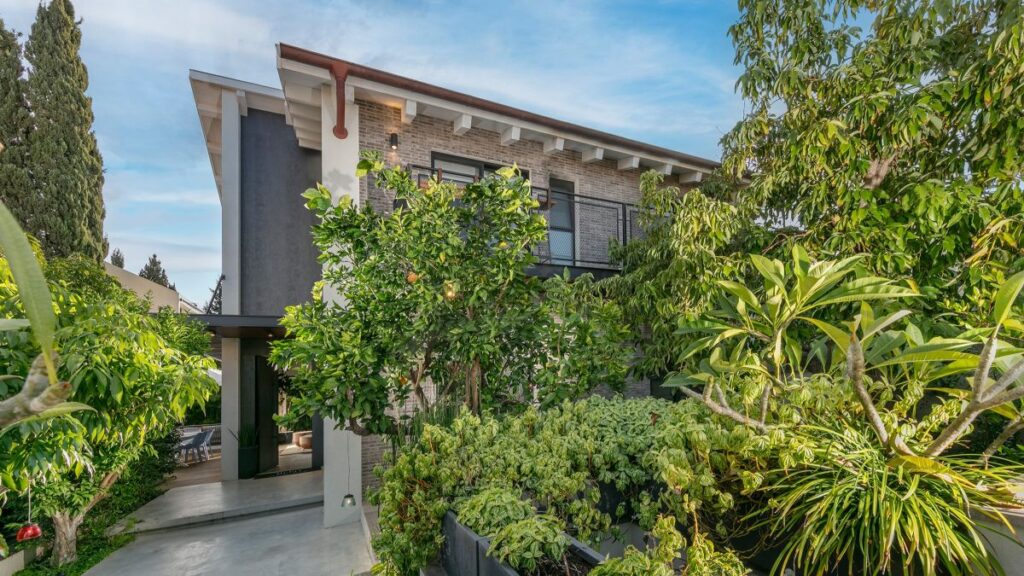 Moderný dom s tehlovou fasádou a terasou obklopený bujnou zeleňou a pestovanými rastlinami.