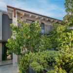 Moderný dom s tehlovou fasádou a terasou obklopený bujnou zeleňou a pestovanými rastlinami.