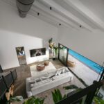 Pohľad zhora na obývaciu izbu s vysokým bielym stropom, šedou sedačkou a výhľadom na bazén.