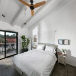 Svetlá spálňa s bielym trámovým stropom, svetlou posteľou a prístupom na balkón.