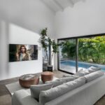 Svetlá obývačka s vysokým stropom, šedou sedačkou, drevenými stolíkmi a výhľadom cez veľké okná na bazén a zeleň.