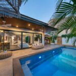 Nočný pohľad na dom s osvetleným bazénom, terasou s ratanovým nábytkom a tropickou zeleňou.