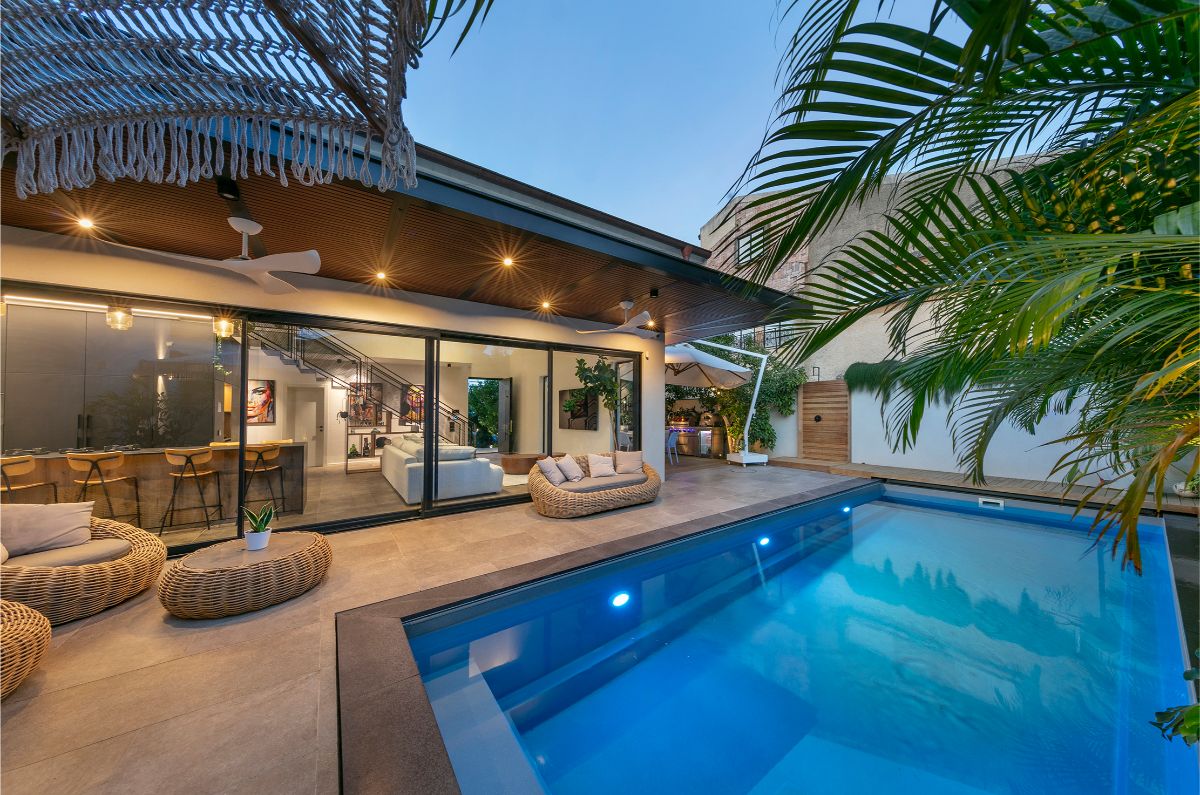 Nočný pohľad na dom s osvetleným bazénom, terasou s ratanovým nábytkom a tropickou zeleňou.