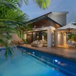 Osvetlený bazén s terasou, jedálenským kútom pod slnečníkom a palmami.