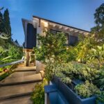 Osvetlený vstup do domu s betónovým chodníkom, tmavou fasádou a bujnou zeleňou.