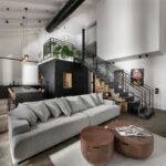 Otvorený priestor s vysokým bielym stropom, šedou sedačkou, čiernym kovovým schodiskom a čiernou kuchynskou linkou v pozadí.