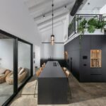 Interiér s vysokým bielym stropom a trámami, čiernou kuchynskou linkou a dlhým ostrovčekom smerujúcim k presklenej stene s terasou.