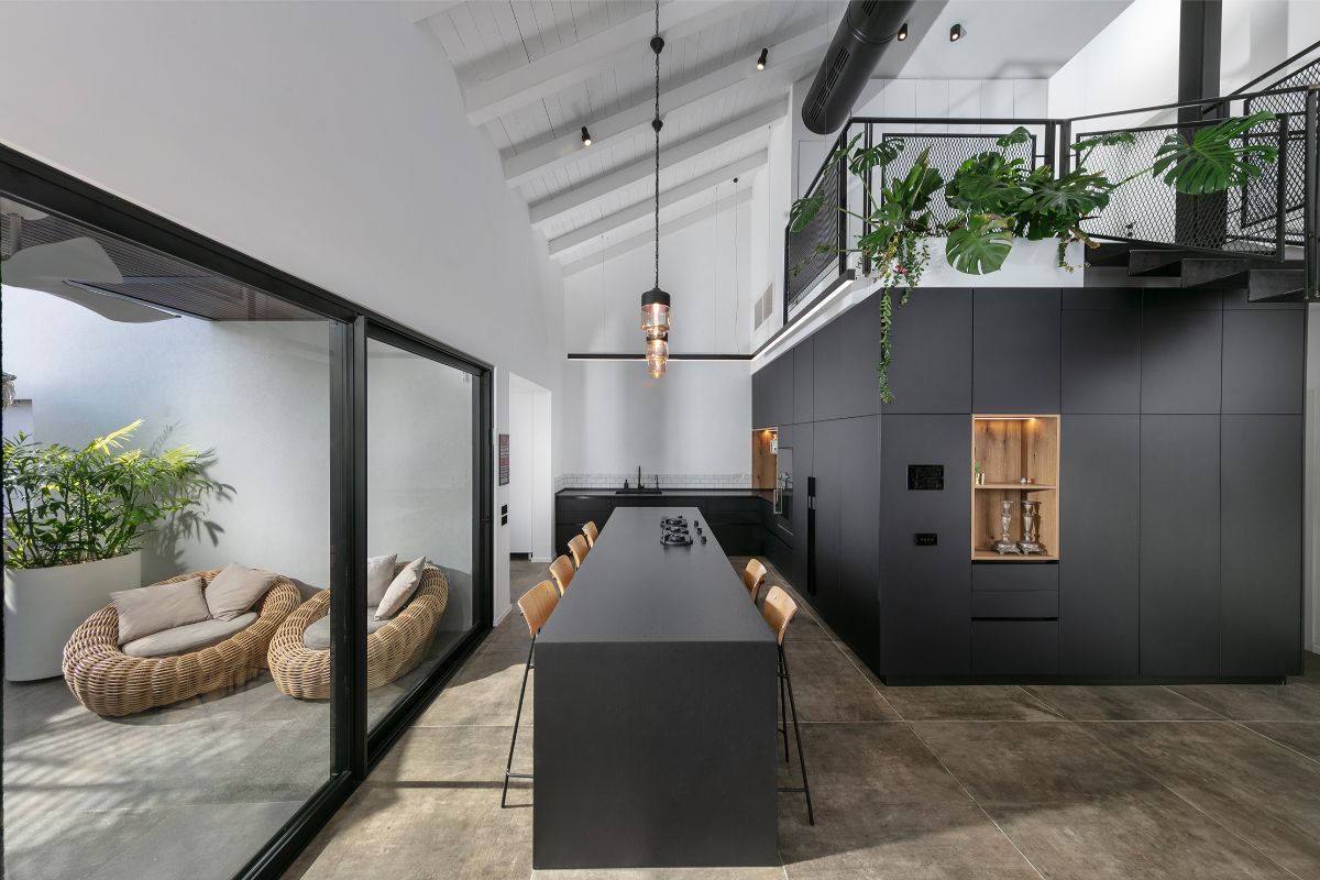 Interiér s vysokým bielym stropom a trámami, čiernou kuchynskou linkou a dlhým ostrovčekom smerujúcim k presklenej stene s terasou.