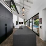 Dlhá čierna kuchynská linka s ostrovčekom a stoličkami pod bielym trámovým šikmým stropom a závesnými svietidlami.