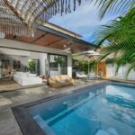 Vonkajší bazén s ležadlami pod tkaným slnečníkom vedľa moderného domu.