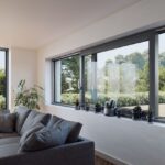 Svetlá obývacia izba s veľkou sivou sedačkou a tmavošedým posuvným oknom s výhľadom do zelene.