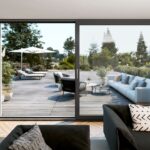 Moderná obývacia izba s čiernou sedačkou a veľkým posuvným oknom vedúcim na drevenú terasu.