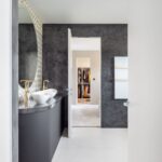 Moderná kúpeľňa s tmavou stierkou, oblé biele umývadlá so zlatými batériami a minimalistické zrkadlo s geometrickým vzorom.