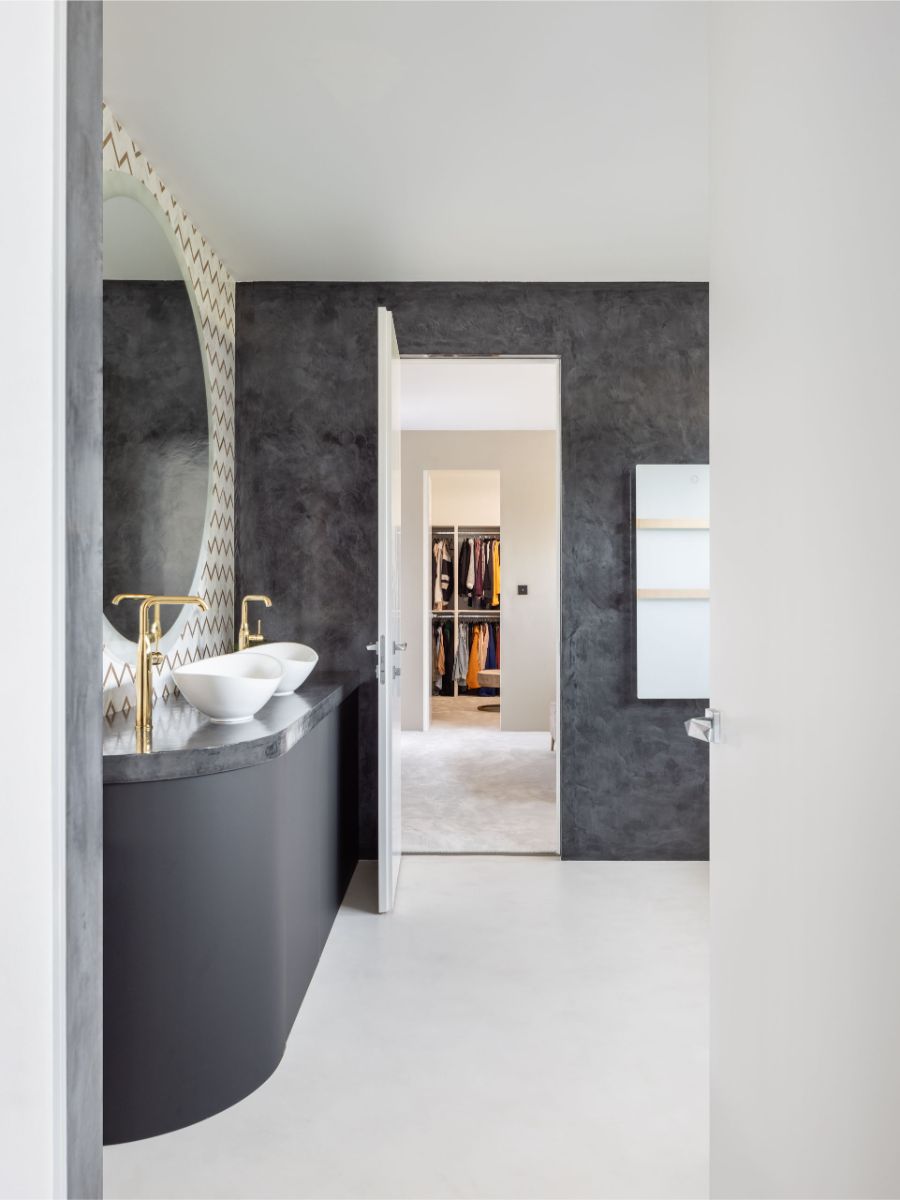 Moderná kúpeľňa s tmavou stierkou, oblé biele umývadlá so zlatými batériami a minimalistické zrkadlo s geometrickým vzorom.