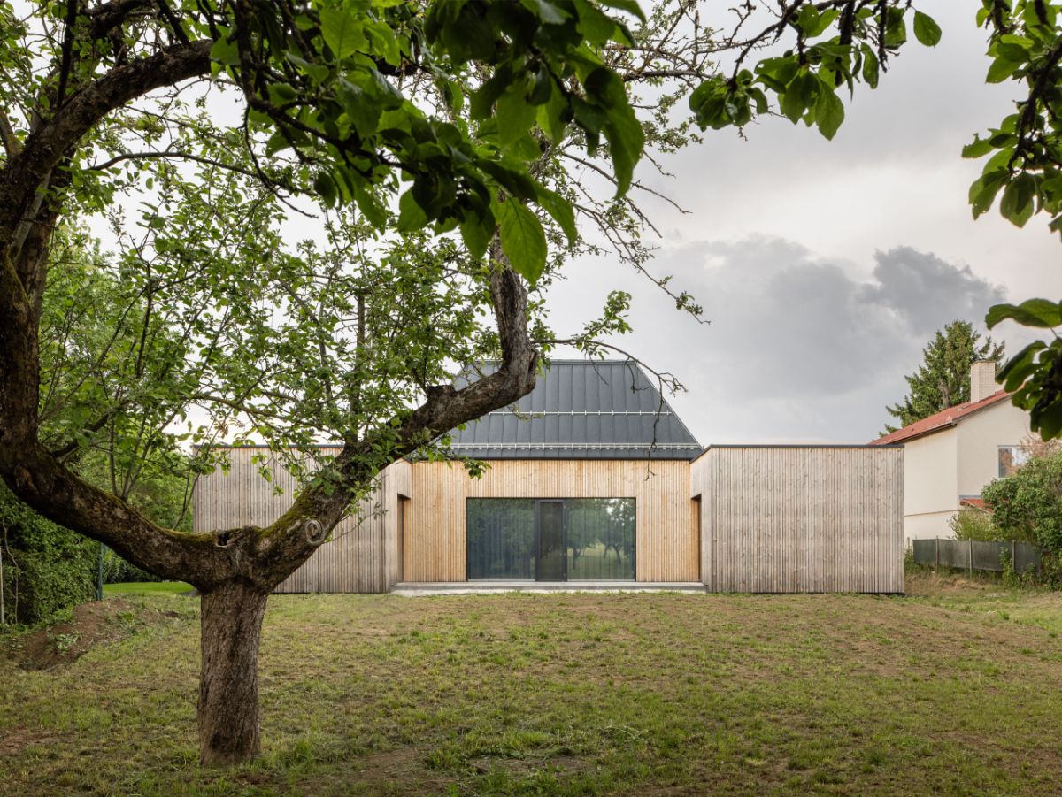 fasáda z prírodného dreva, tmavá sedlová strecha átriového domu a zatrávnená záhrada s ovocným stromom.