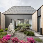 Pohľad cez átrium s chodníkom z dlažby a minimalistickými záhonmi na obytnú časť domu s valbovou strechou.