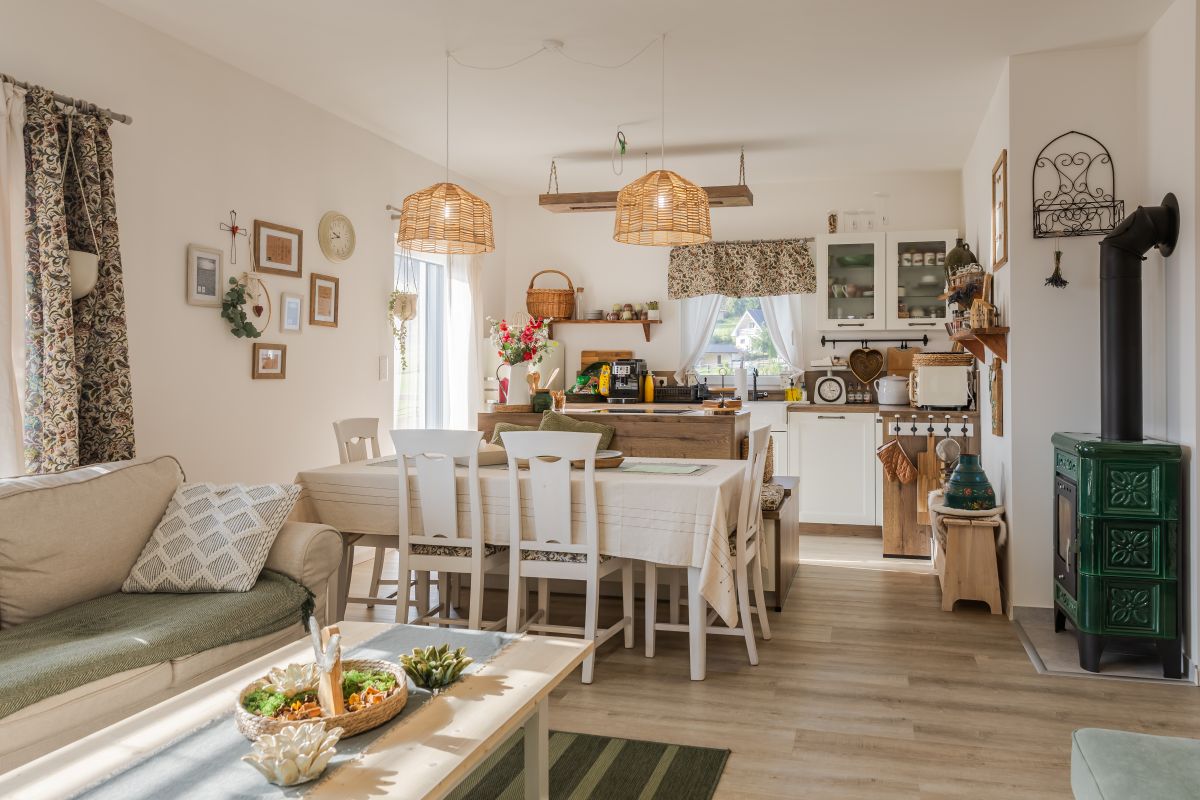 Otvorená bielo-drevená kuchyňa spojená s obývačkou v rustikálnom štýle, s bielym jedálenským stolom a zelenou kachľovou pecou.
