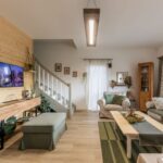 Svetlá obývačka s drevenou stenou, béžovou sedačkou a zeleným otomanom, so schodiskom a rustikálnym dreveným nábytkom