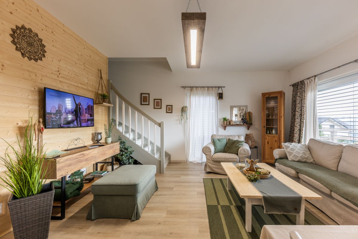 Svetlá obývačka s drevenou stenou, béžovou sedačkou a zeleným otomanom, so schodiskom a rustikálnym dreveným nábytkom