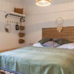 Spálňa v prírodnom štýle s posteľou na drevených paletách, prúteným čelom a tienidlom, so zelenou prešívanou prikrývkou.