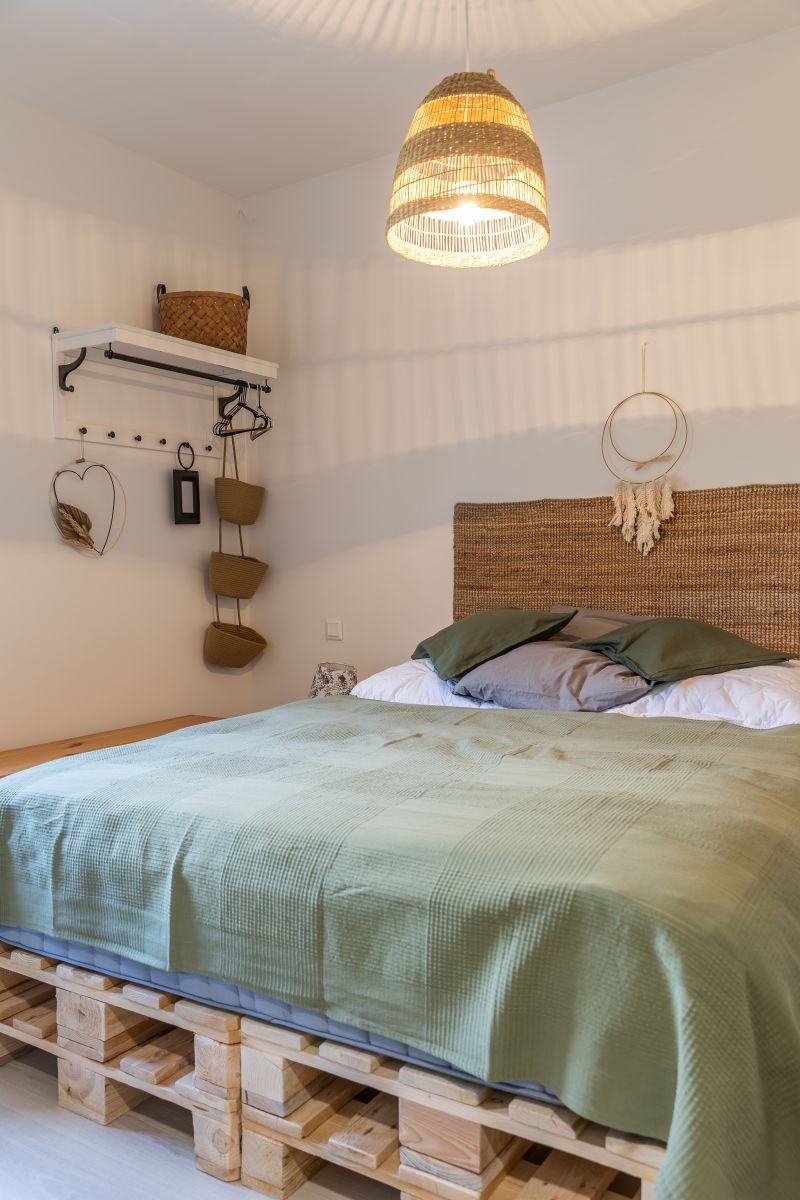 Spálňa v prírodnom štýle s posteľou na drevených paletách, prúteným čelom a tienidlom, so zelenou prešívanou prikrývkou.