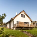 Biely rodinný dom s hnedou strechou a priestrannou drevenou terasou, stojaci na svahu s trávnatým pozemkom v dedinskom prostredí.