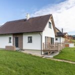 Moderný biely dom s hnedou strechou a hnedou drevenou terasou, obklopený trávou na miernom svahu s kamenným chodníkom.