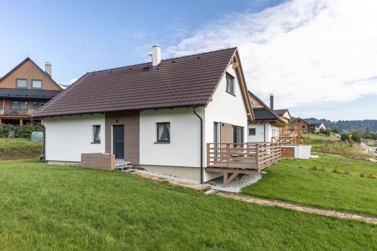 Moderný biely dom s hnedou strechou a hnedou drevenou terasou, obklopený trávou na miernom svahu s kamenným chodníkom.