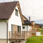 Bočný pohľad na moderný biely dom s hnedou strechou a drevenou terasou s ratanovým nábytkom.