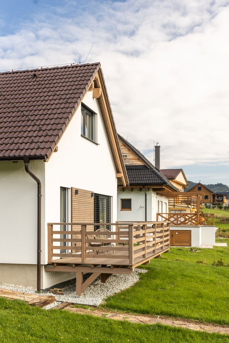 Bočný pohľad na moderný biely dom s hnedou strechou a drevenou terasou s ratanovým nábytkom.