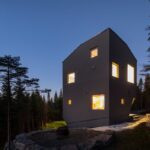 Tmavý dom obklopený hustým ihličnatým lesom a rozsiahlym výhľadom na hory.