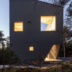 Exteriér moderného domu so asymetrickou strechou, teplým osvetlením a s hustým ihličnatým lesom pri súmraku.