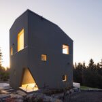 Moderný tmavý dom zasadený hlboko v lese s drevenými rámami okien a svetlým interiérom.