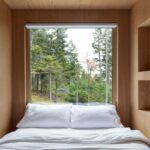Posteľ obklopená dreveným obložením, veľké okno s výhľadom na hustý ihličnatý les, biela posteľná bielizeň.
