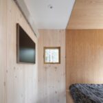 Časť spálne s obkladom zo svetlého smrekového dreva, šedá podlaha a televízia na stene.