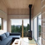 Obývacia izba s dreveným interiérom, tmavosivou sedacou súpravou a čiernym krbom pri veľkom okne do lesa.