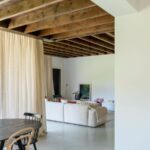 Prepojený priestor jedálne a obývačky s dreveným trámovým stropom, bielym gaučom a veľkým oknom.