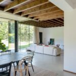 Prepojený priestor jedálne a obývačky s dreveným trámovým stropom, bielym gaučom a veľkým oknom.