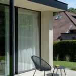 Čierna drôtená záhradná stolička a stolík na terase pri veľkej presklenej stene s bielym závesom.