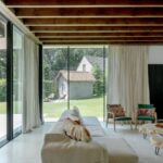 Presvetlená obývačka s béžovou sedačkou, zelenými kreslami a širokými drevenými trámami nad sklenou stenou.