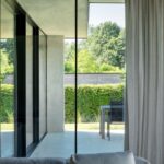 Presklený roh modernej obývačky s terasou, záhradným sedením a vysokým živým plotom.