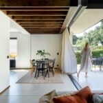 Žena v bielych šatách pri závese medzi jedálňou s okrúhlym čiernym stolom a vonkajšou terasou.